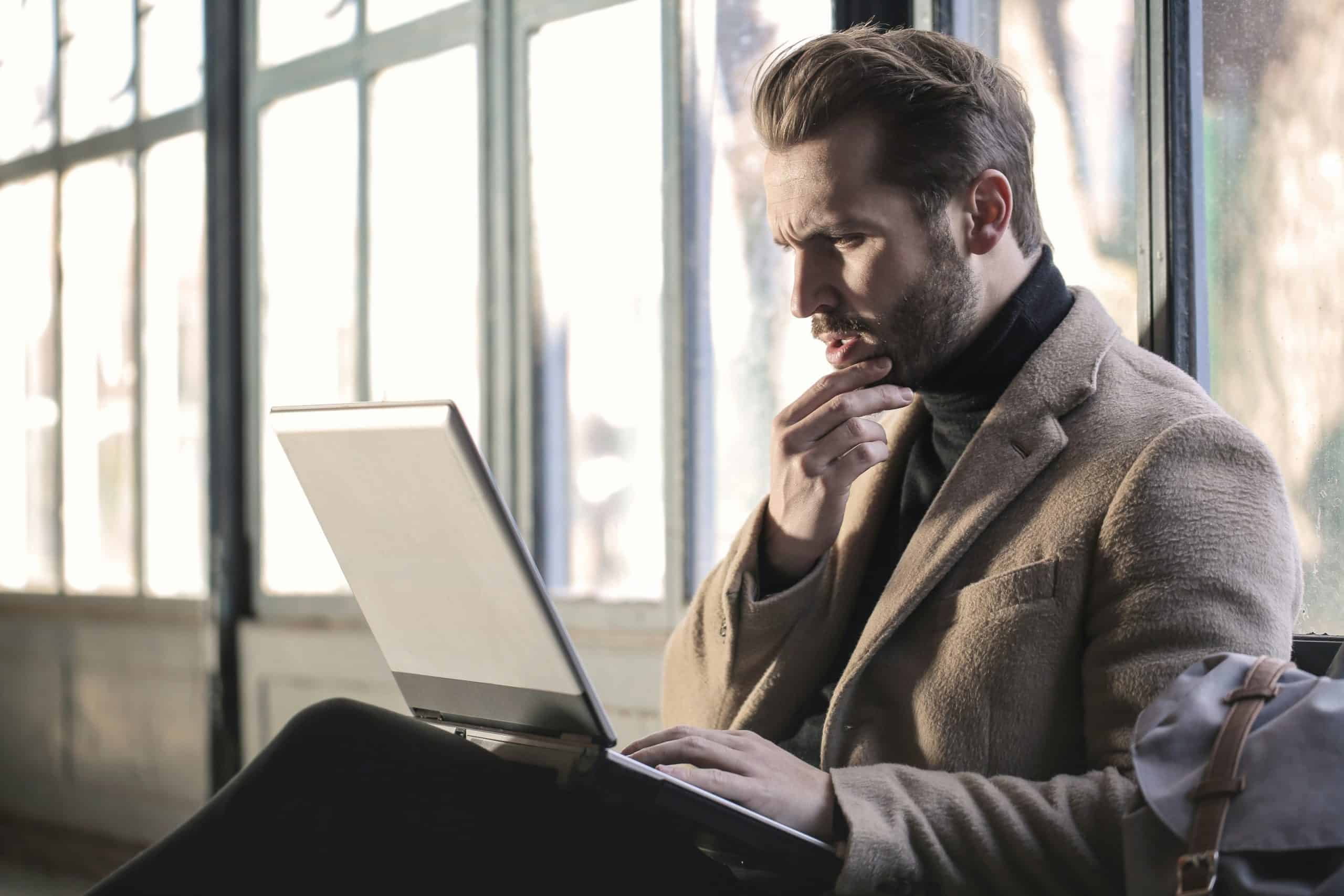 En mann i frakk sitter foran en laptop og vurderer sine valg før han søker om bedriftslån.